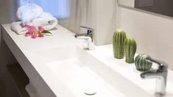 Une salle de bain avec un lavabo blanc, deux robinets, une serviette, une fleur rose et deux cactus verts.