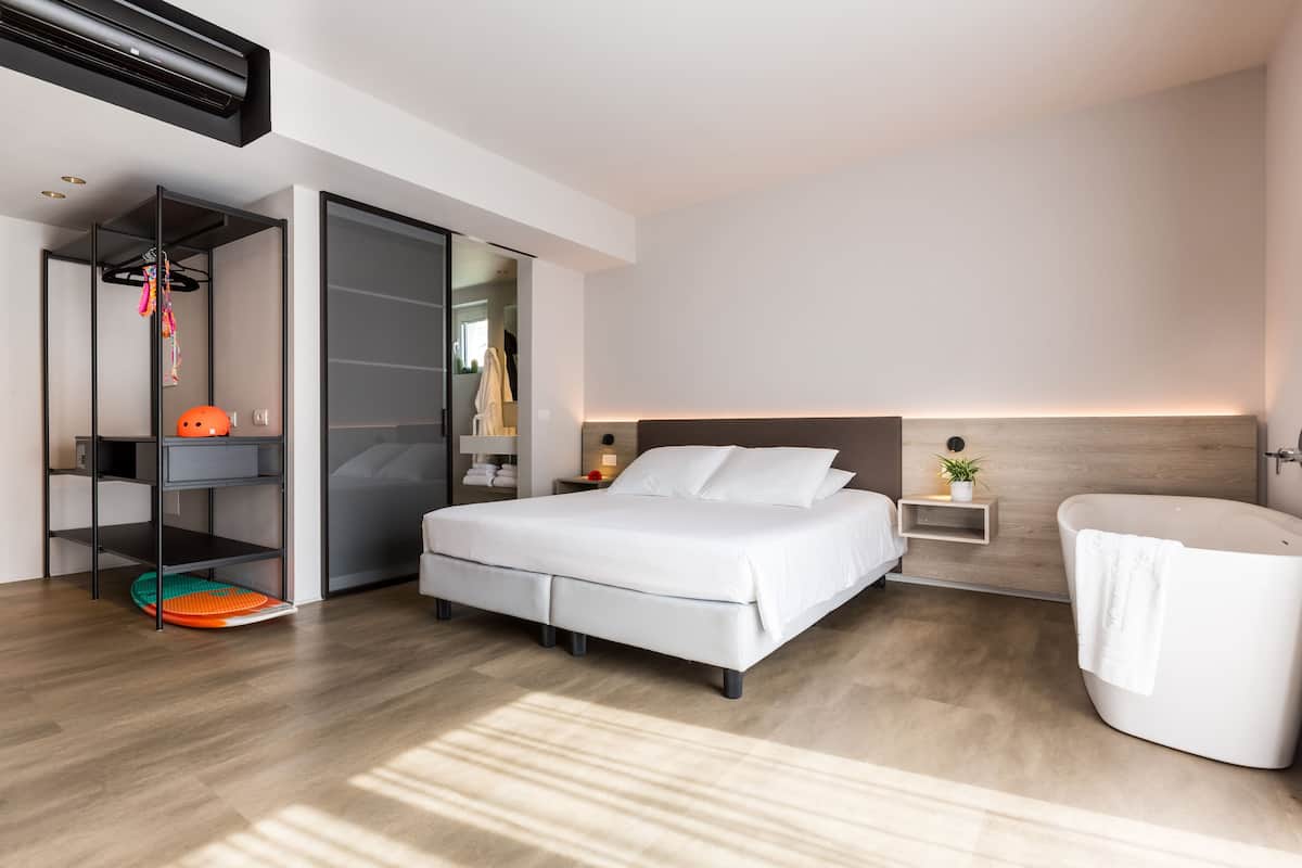Moderni hotellihuone, jossa on suuri sänky, sisäänrakennettu vaatekaappi, yöpöytä lampulla sekä erillinen kylpyhuone, jossa on kylpyamme.