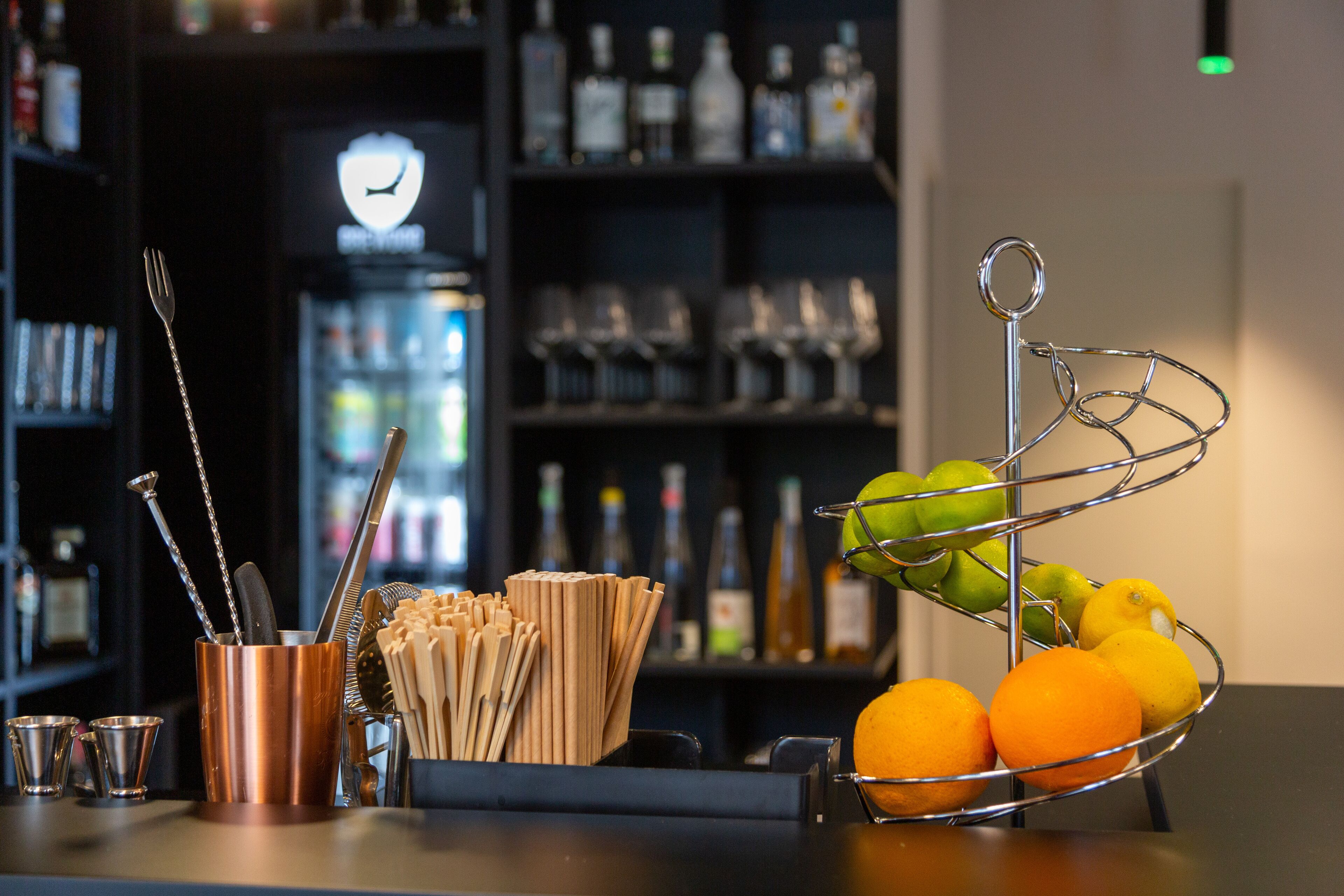 Una zona de bar con una cesta de metal llena de naranjas y limones, un vaso de cobre con utensilios, y un fondo con estantes de botellas y vasos.
