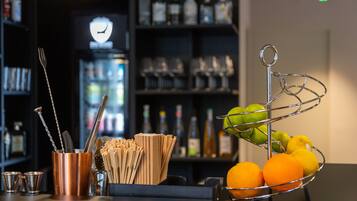 Una zona de bar con una cesta de metal llena de naranjas y limones, un vaso de cobre con utensilios, y un fondo con estantes de botellas y vasos.