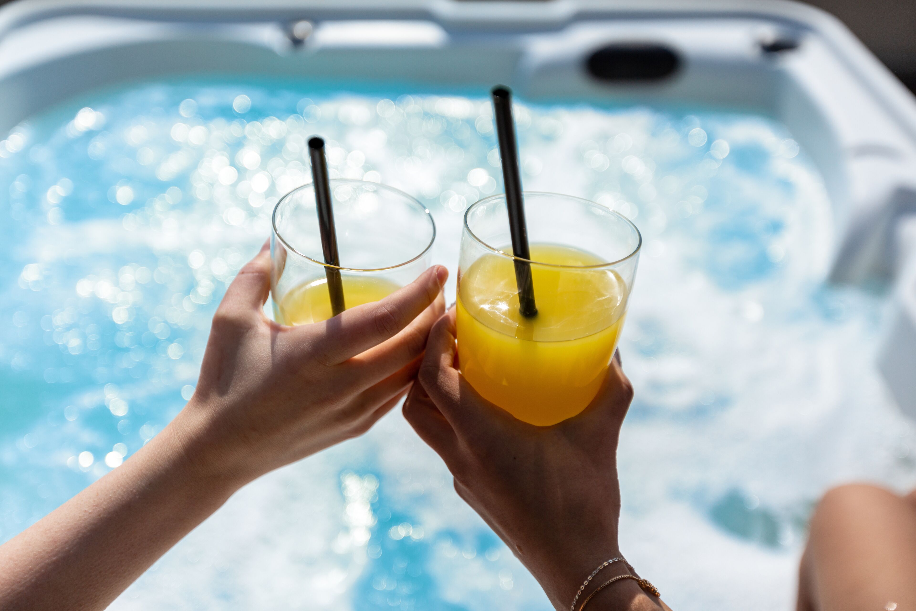 Dos manos sosteniendo vasos con líquido anaranjado y pajitas, con una piscina al fondo.