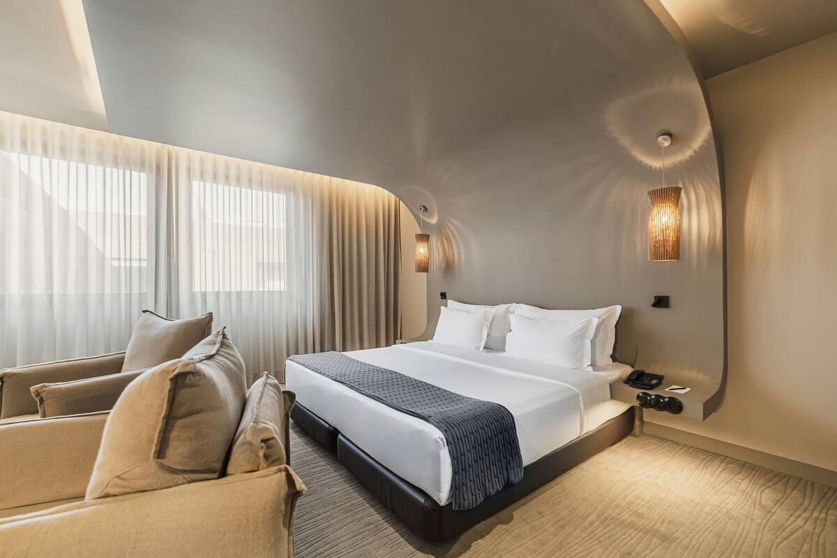 A modern hotel room with a large bed, a sofa, a desk with a phone, and a wall-mounted lamp.