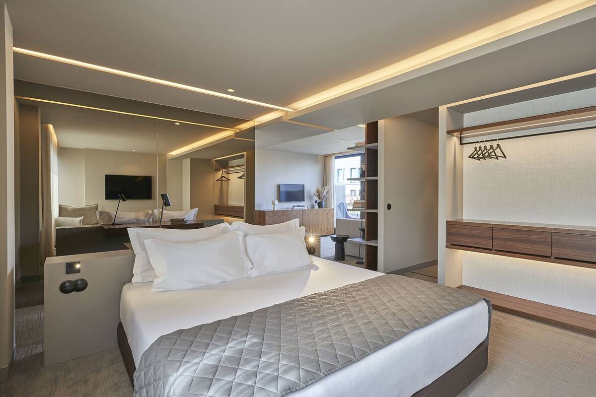 A modern hotel room with a large bed, a bedside table, a TV, a mirror, and a wardrobe.