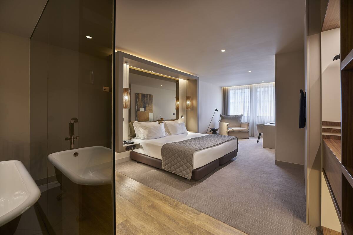 A modern hotel room with a large bed, a sitting area, a bathtub, and a large mirror.