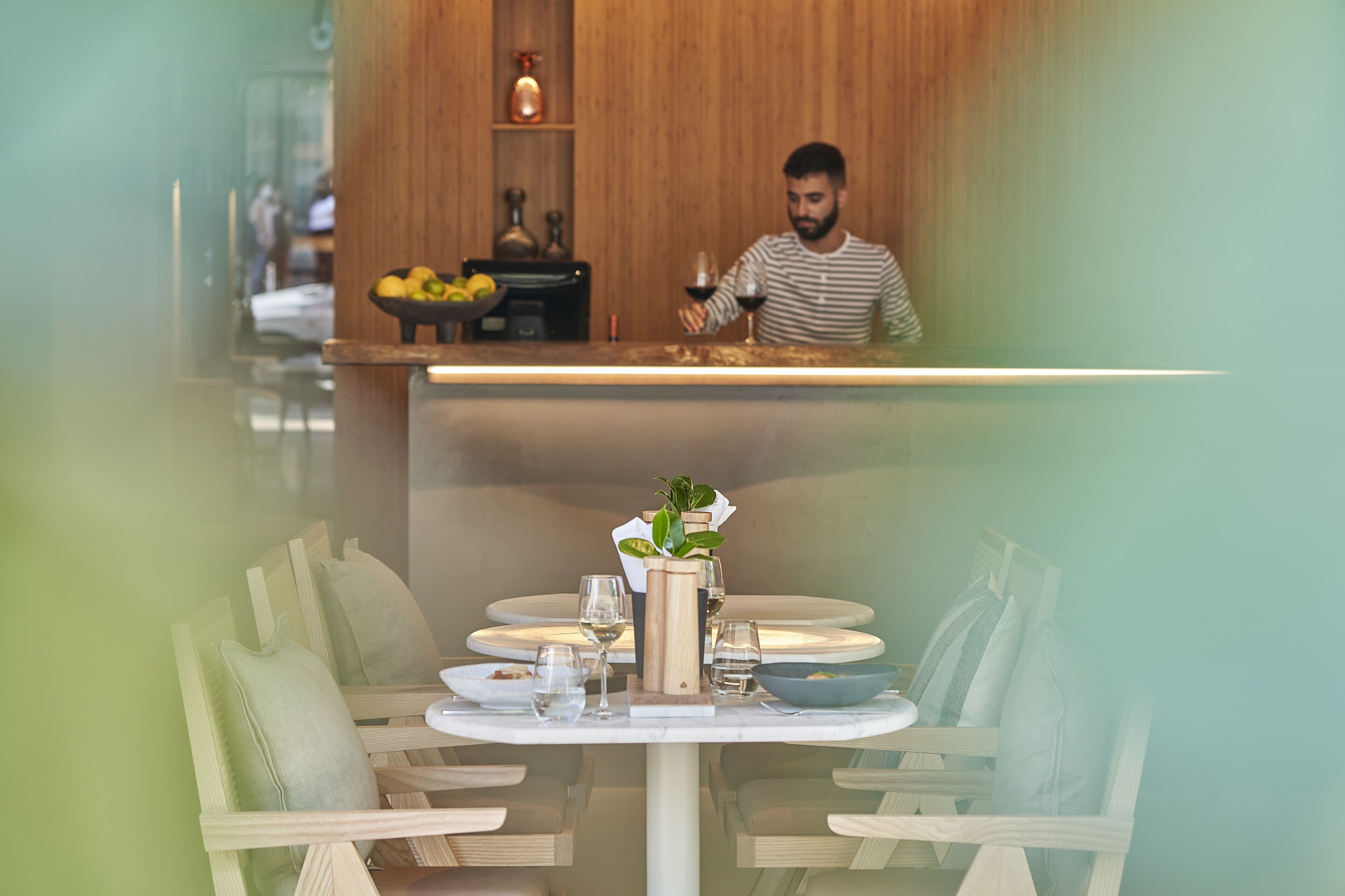 Un restaurant avec un décor intérieur en bois, un comptoir derrière lequel se trouve une personne, et une table dressée avec des assiettes, des verres et un vase rempli de fleurs.