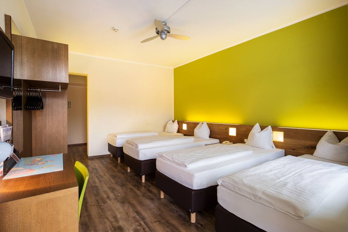 Habitación de hotel con cuatro camas individuales, un escritorio con silla, un ventilador de techo y un armario de madera.