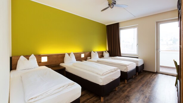 A hotel room with three beds, wooden flooring, a ceiling fan, and a large window. - Basic Hotel Innsbruck (Innsbruck)