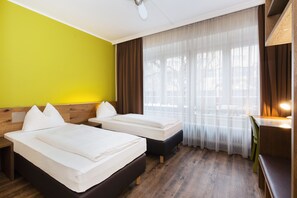 A hotel room with two beds, a desk, and a large window with sheer curtains.