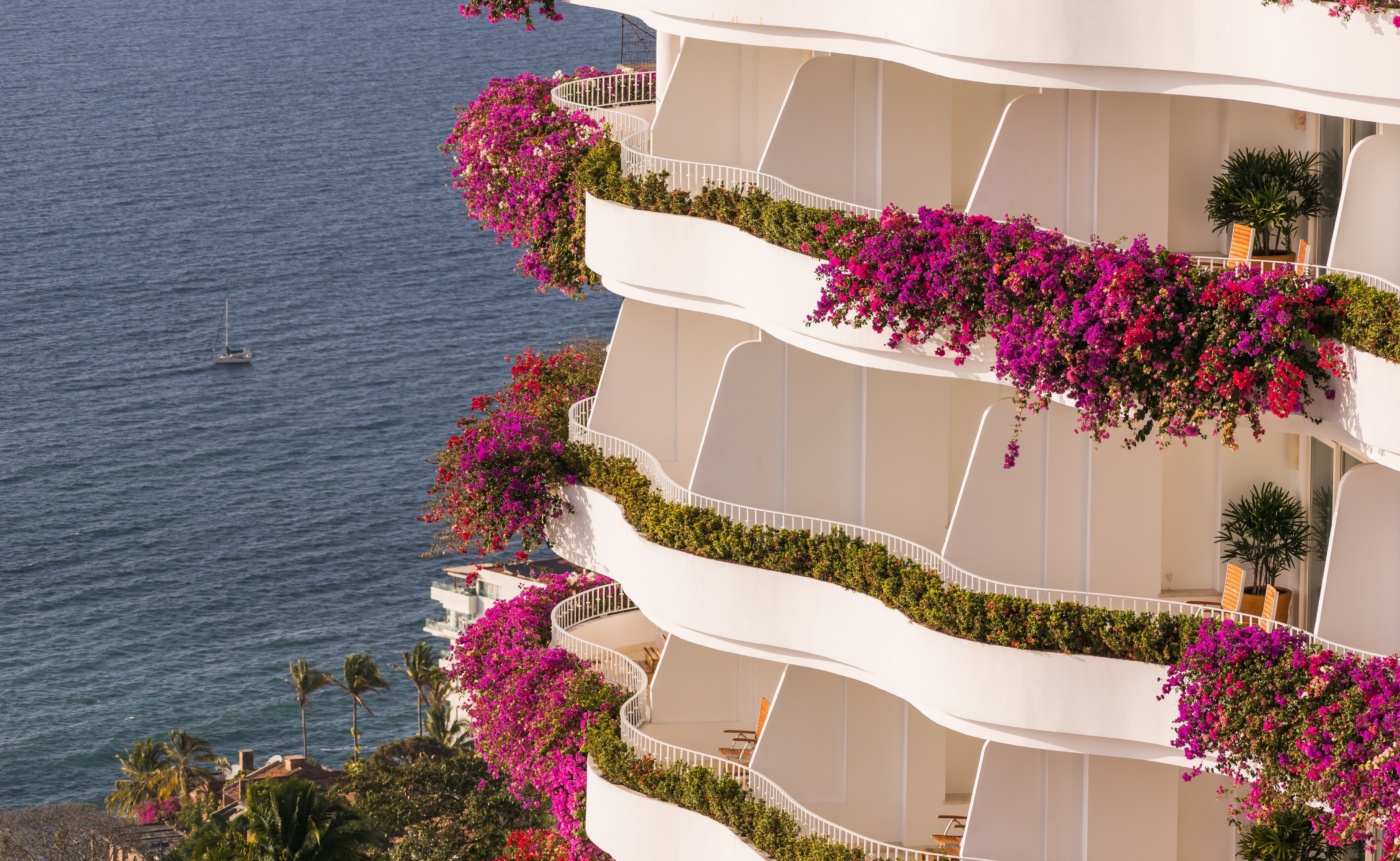 Et hotel med flere etager og balkoner pyntet med livlige pink blomster, med udsigt over et roligt hav.