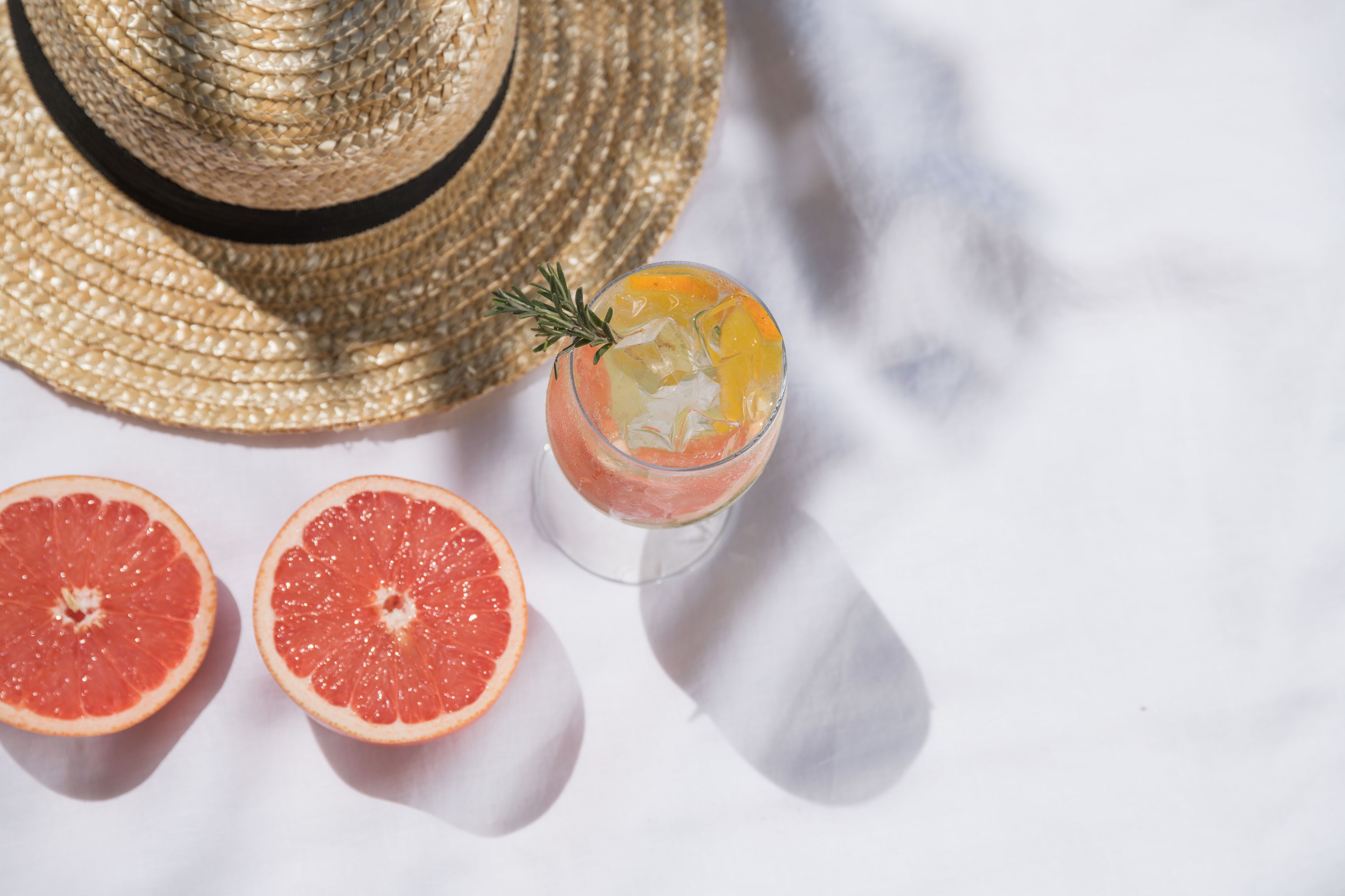Un sombrero de paja, dos mitades de pomelo y un vaso con una bebida y una ramita de romero.