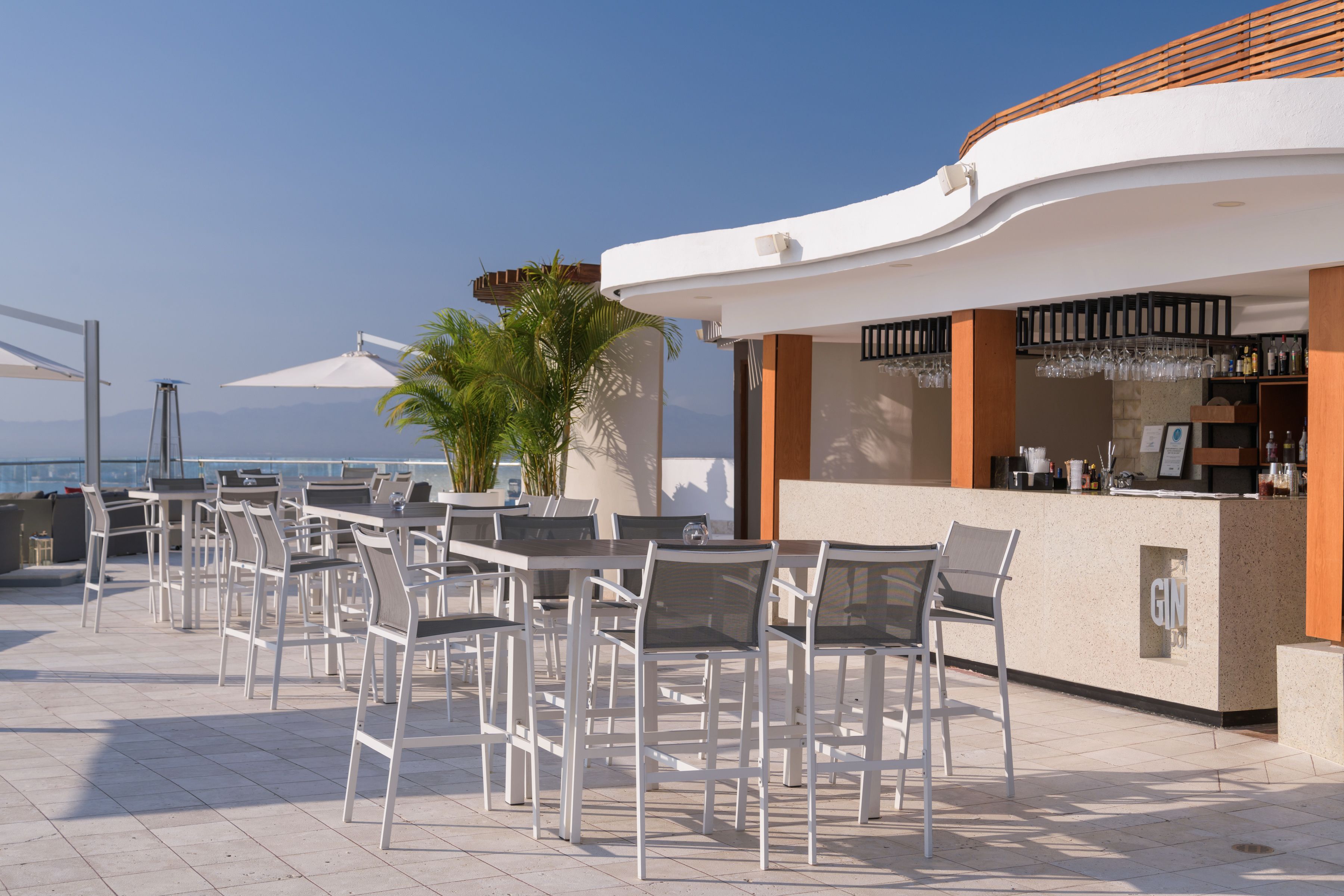 An outdoor seating area with tables and chairs, a bar counter, and a view of the sea.