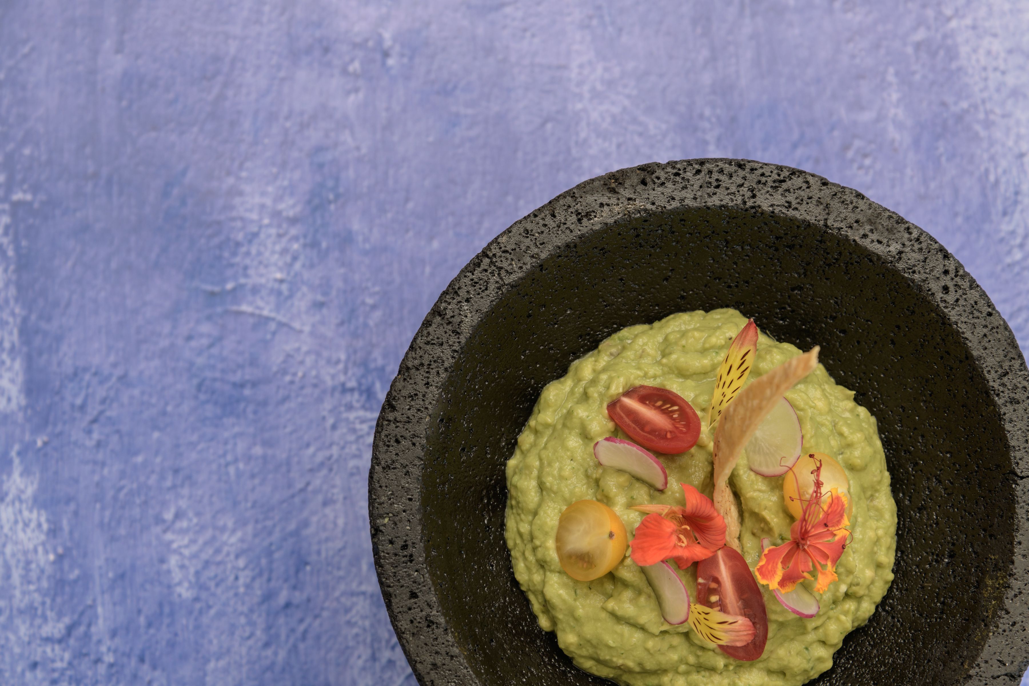 A bowl of green dip garnished with slices of tomato, radish, and edible flowers on a textured blue background.