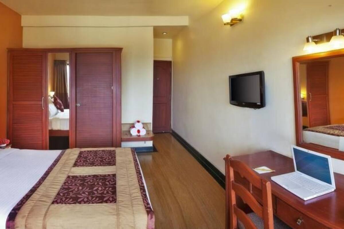 Quarto de hotel com cama, mesa de cabeceira, escrivaninha, cadeira e televisão.