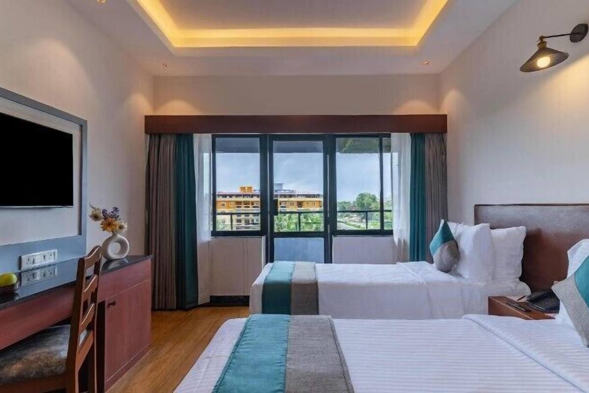 Quarto de hotel com duas camas, televisão, escrivaninha e uma grande janela com vista para edifícios.
