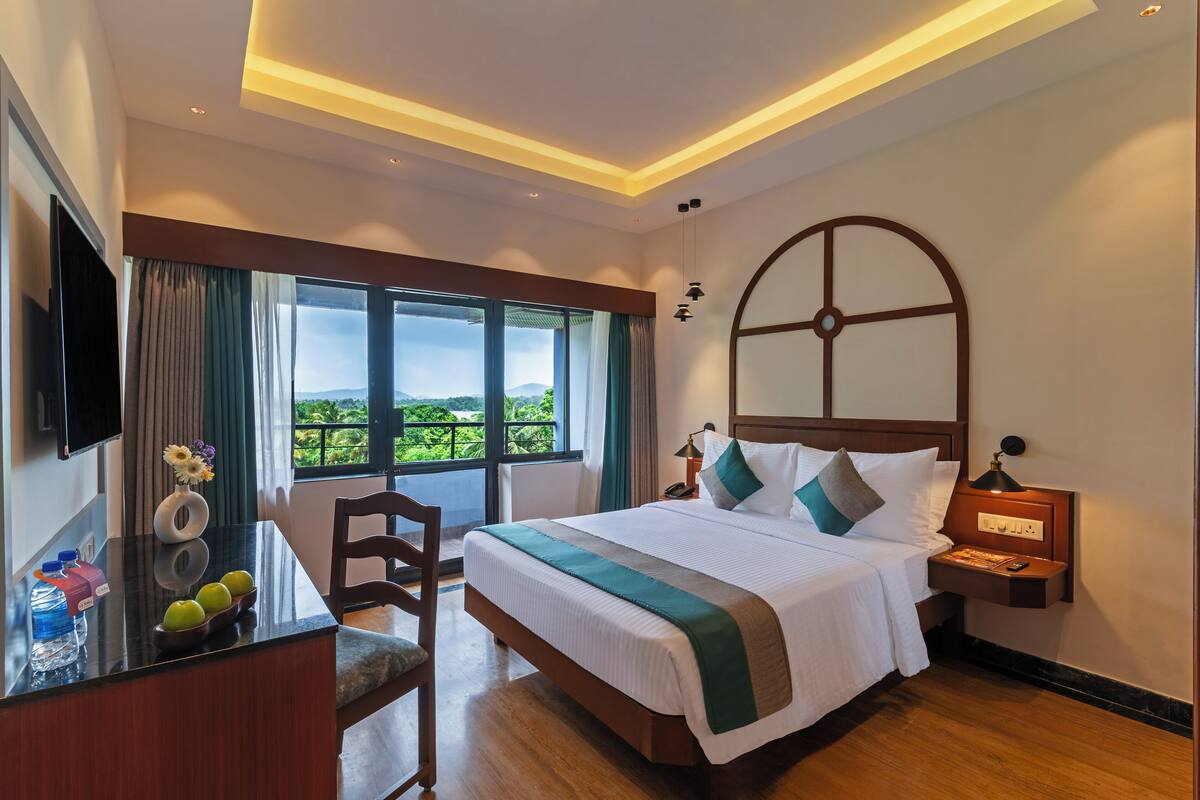 Quarto de hotel com uma cama grande, uma mesa com cadeira, uma TV e uma janela com vista para áreas verdes.