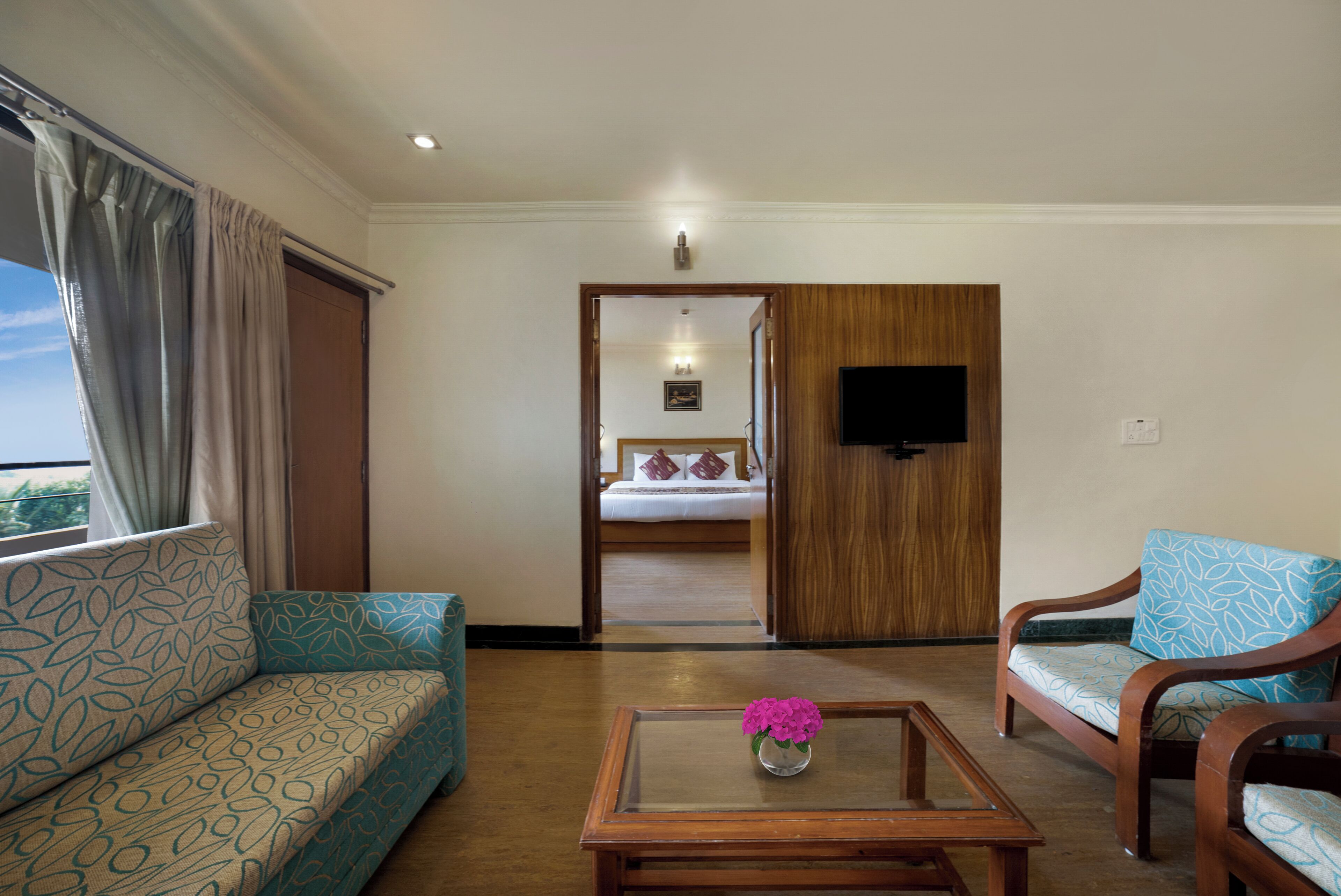 A hotel room with a sofa, a coffee table with flowers, a TV, and a bed with pillows.
