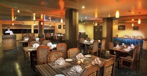 A restaurant with multiple dining tables, each set with plates, cutlery, and napkins. There are wooden chairs with intricate designs. A counter with a person behind it is visible, suggesting a service area. - Sterling Karwar (Karwar)