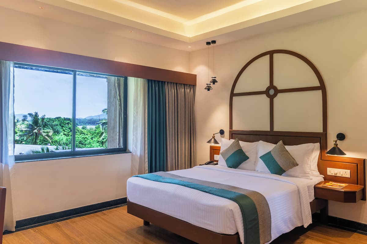 Quarto de hotel com uma cama grande, uma mesa de cabeceira com abajur, janela com cortinas e vista para o verde lá fora.