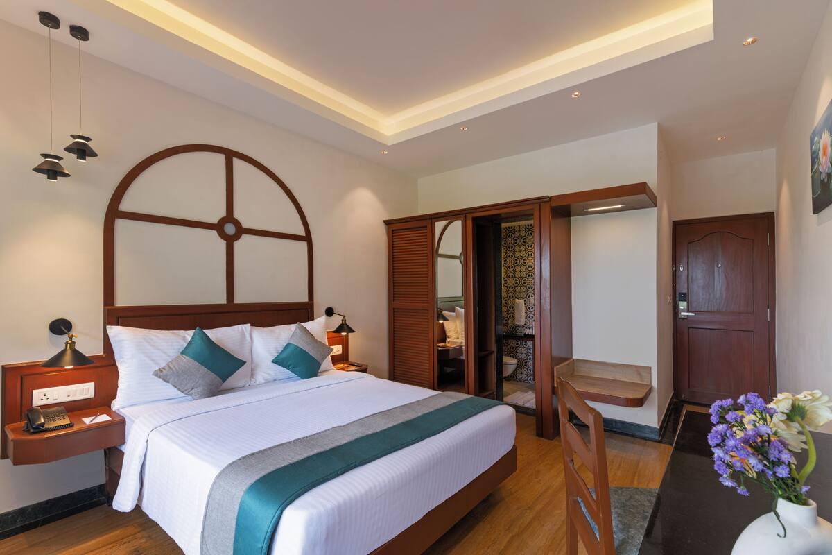 Quarto de hotel com uma cama grande, mesa de cabeceira com abajur, uma cadeira de madeira, uma pequena mesa com flores e uma porta que dá para um banheiro.