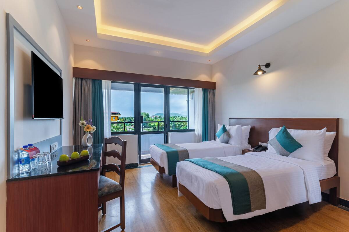 Quarto de hotel com duas camas, uma escrivaninha com televisão de tela plana, uma cadeira e uma janela grande com vista para área verde.