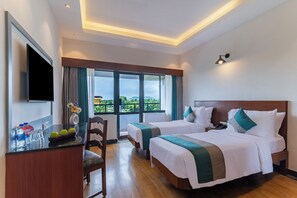 A hotel room with two beds, a desk with a flat-screen TV, a chair, and a large window offering a view of greenery. - Sterling Karwar (Karwar)