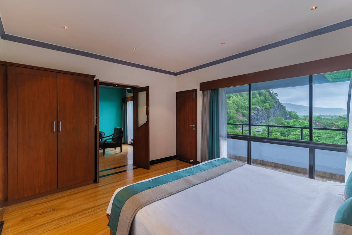Um quarto amplo com uma cama grande, guarda-roupa de madeira e vista para o verde através de uma janela grande.