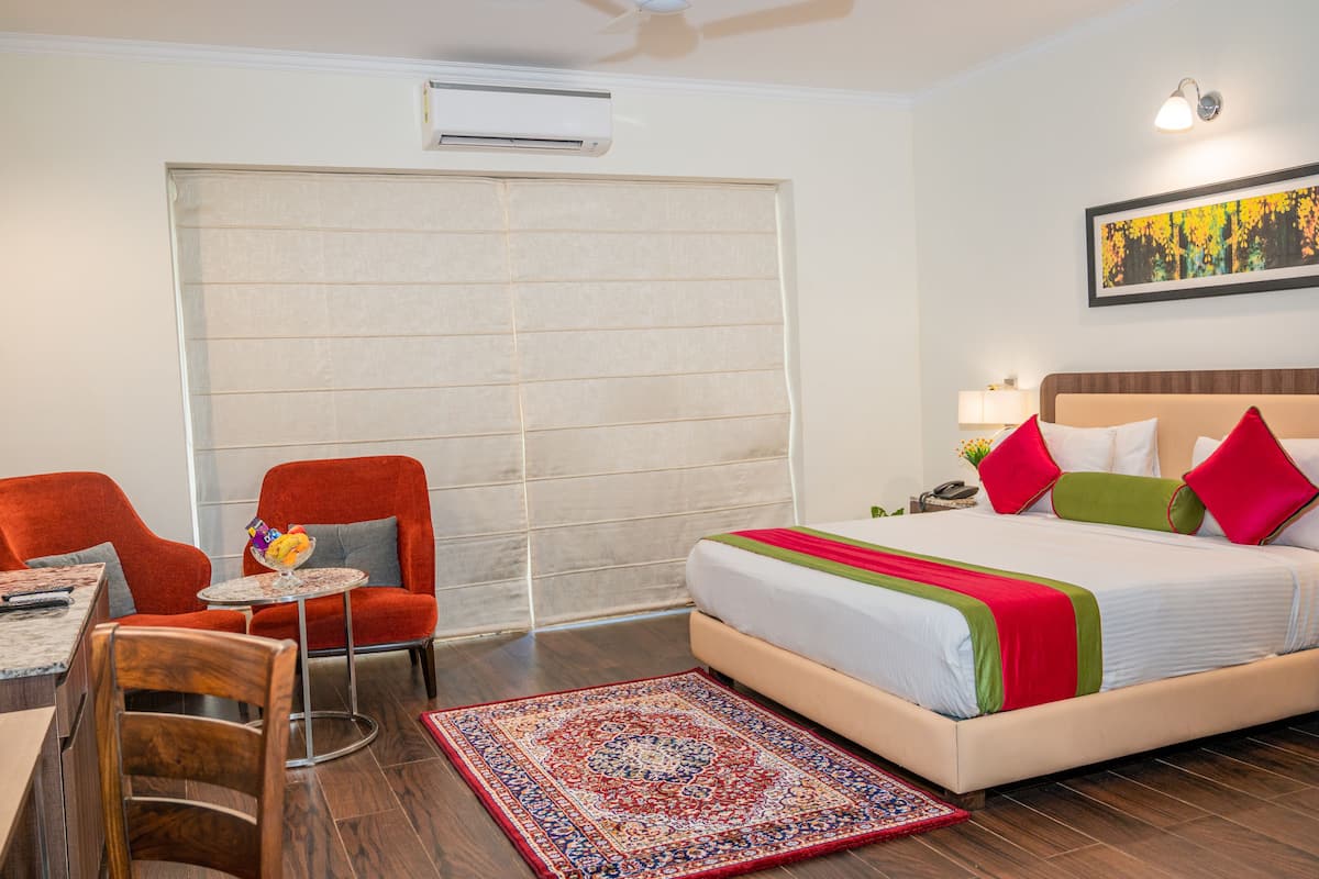 Une chambre d’hôtel avec un grand lit, un fauteuil rouge, une petite table sur laquelle se trouve un saladier de fruits, et une grande fenêtre avec des stores.
