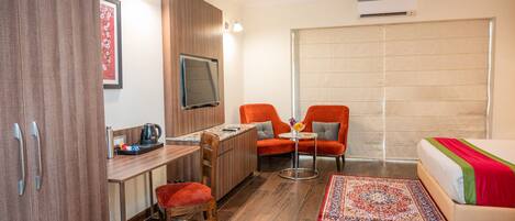 A hotel room with a wooden desk, two orange chairs, a bed with a red and green bedspread, a television, and a ceiling fan.