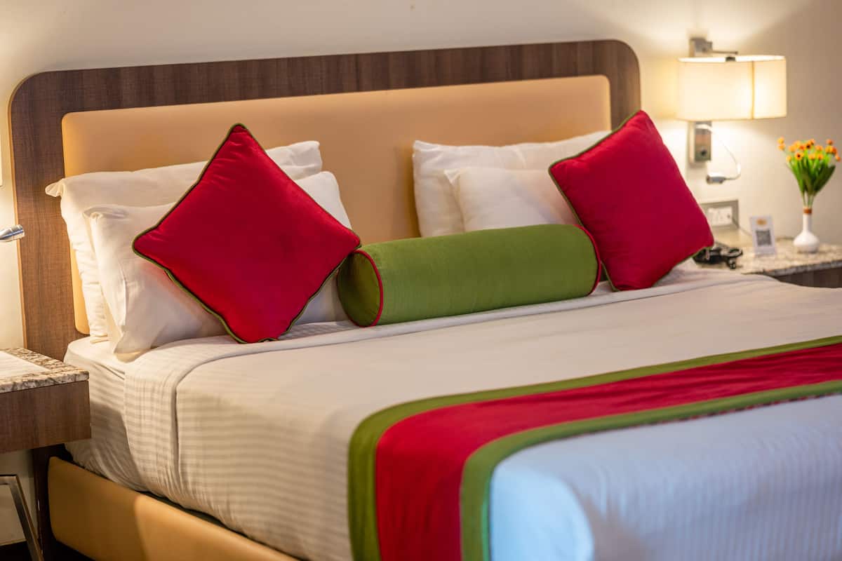 Une chambre d’hôtel avec un lit, des oreillers, une table de chevet, une lampe et un vase rempli de fleurs.