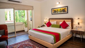 Quarto de hotel com uma cama grande, uma cadeira vermelha, uma mesa pequena, uma mesinha de cabeceira, uma luminária, um quadro emoldurado e uma janela com vista para o verde.