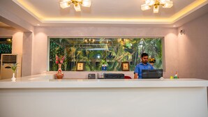 Balcão de recepção com uma grande janela que oferece vista tropical, um ventilador de teto e um certificado emoldurado.