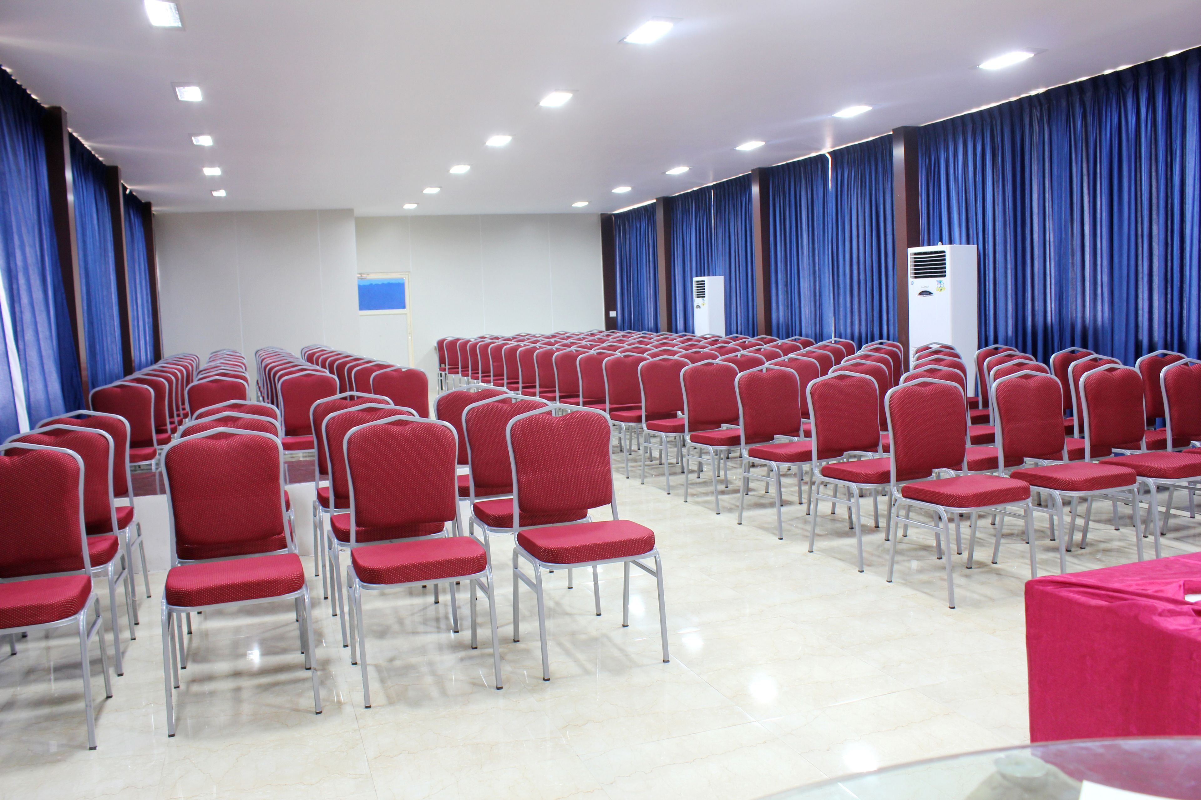 Un salón de conferencias con filas de sillas rojas, un equipo de aire acondicionado grande y cortinas azules.