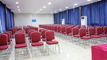 Un salón de conferencias con filas de sillas rojas, un equipo de aire acondicionado grande y cortinas azules.