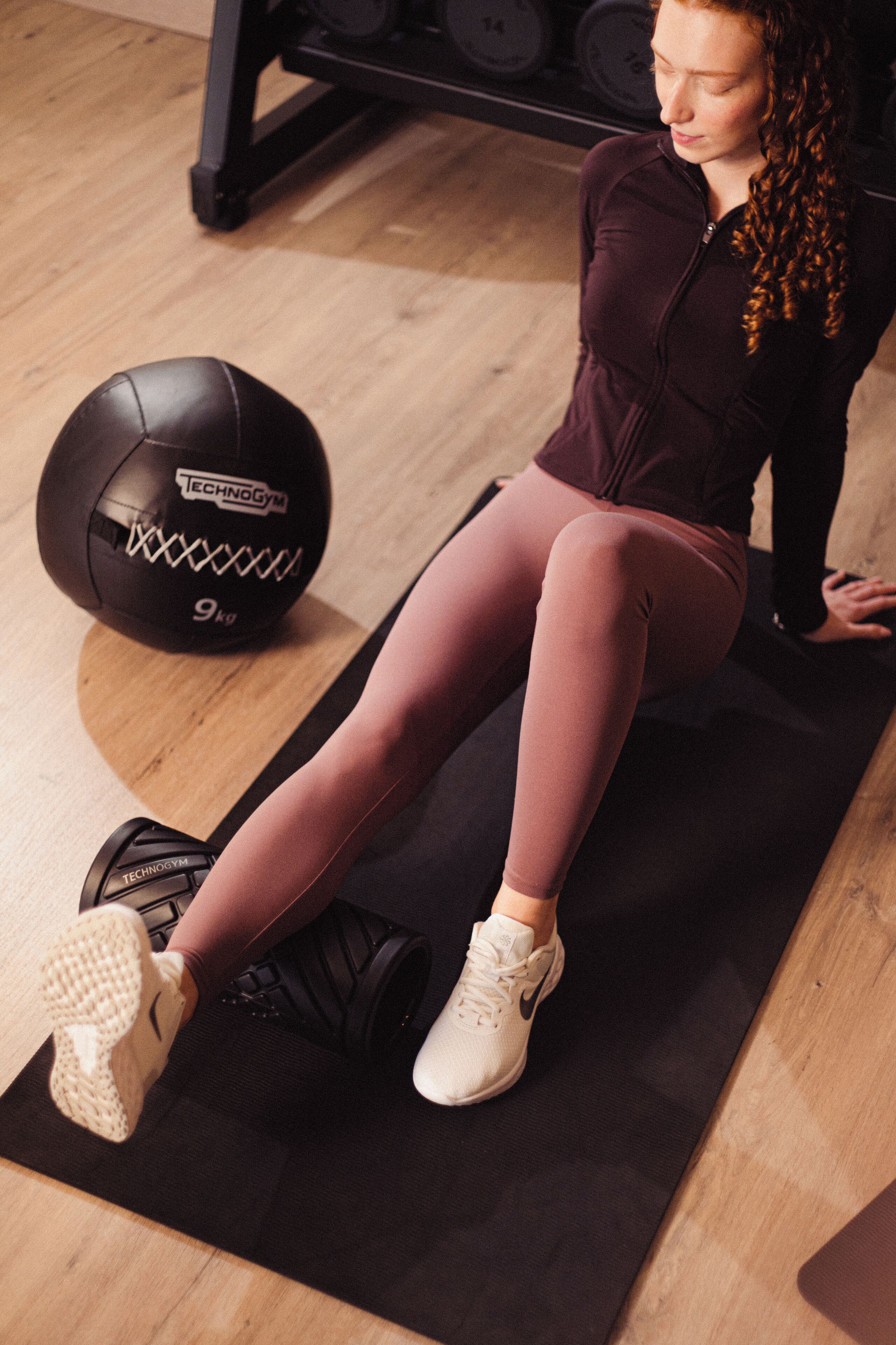 Une personne est assise sur un tapis de sport noir, avec un ballon de gymnastique noir portant les inscriptions « Technogym » et « 9 kg », le tout posé sur un plancher en bois.