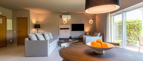 A modern living room with a sofa, a dining table with a bowl of oranges, and a large window with a view of greenery.