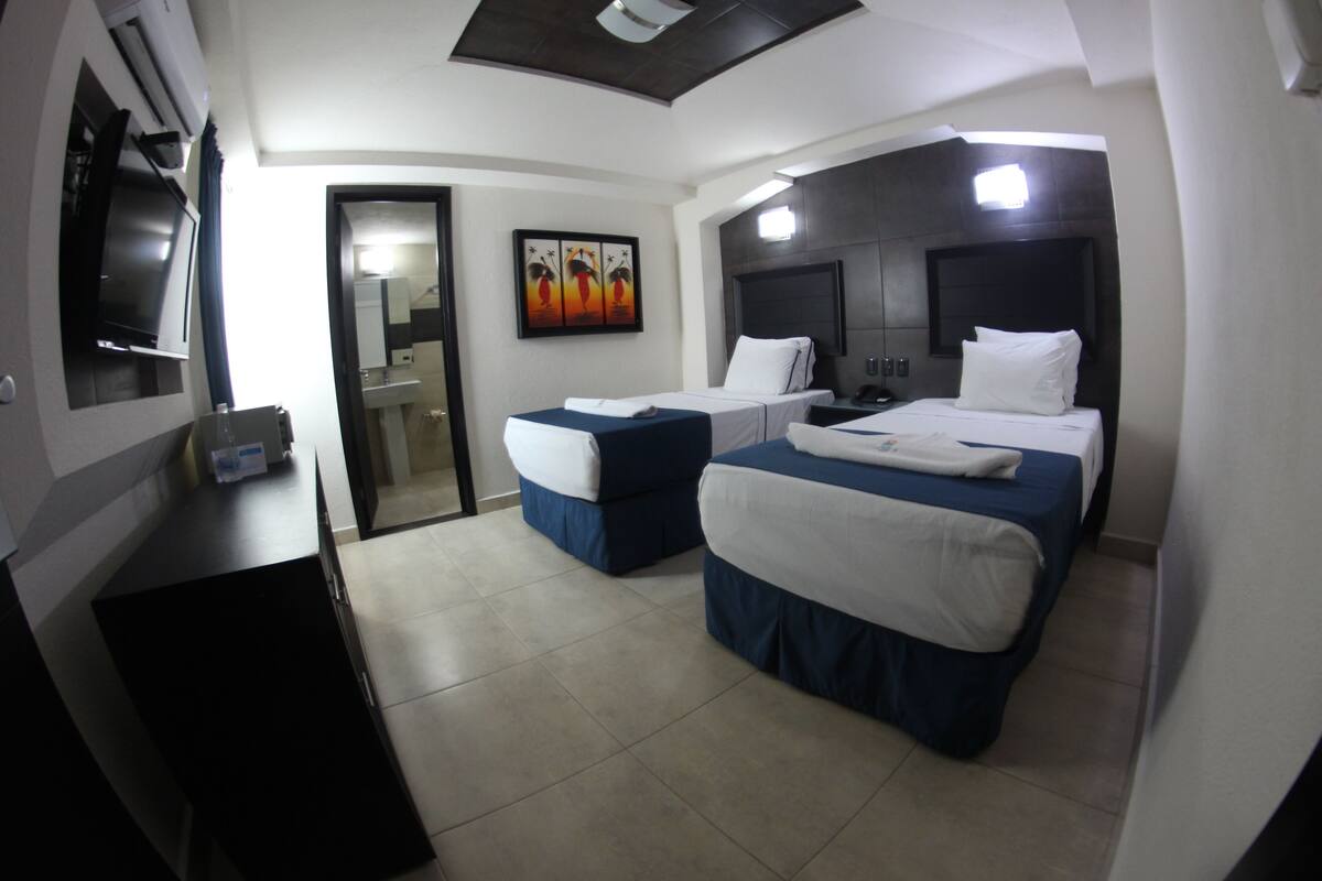 Habitación de hotel con dos camas, televisión, escritorio y baño visible a través de una puerta abierta.