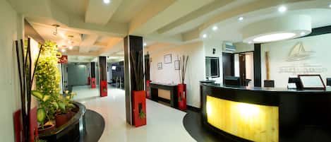 A hotel reception area with a curved reception desk, potted plants, and framed certificates on the wall.