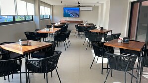 A conference room with multiple tables and chairs, a large flat-screen TV on the wall, and a view of the outdoors through the windows. - Hotel Portonovo Plaza Malecón (Puerto Vallarta)
