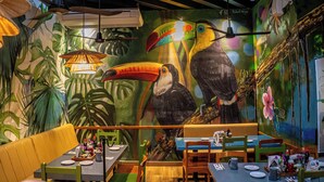 A restaurant interior with tropical murals, wooden furniture, and hanging pendant lights.