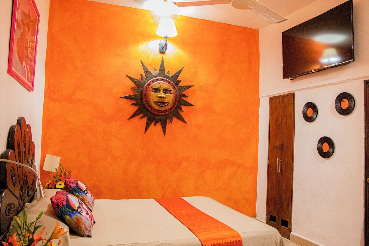 A room with an orange accent wall, a bed with an orange blanket, a television on the wall, and a door with a wooden frame.