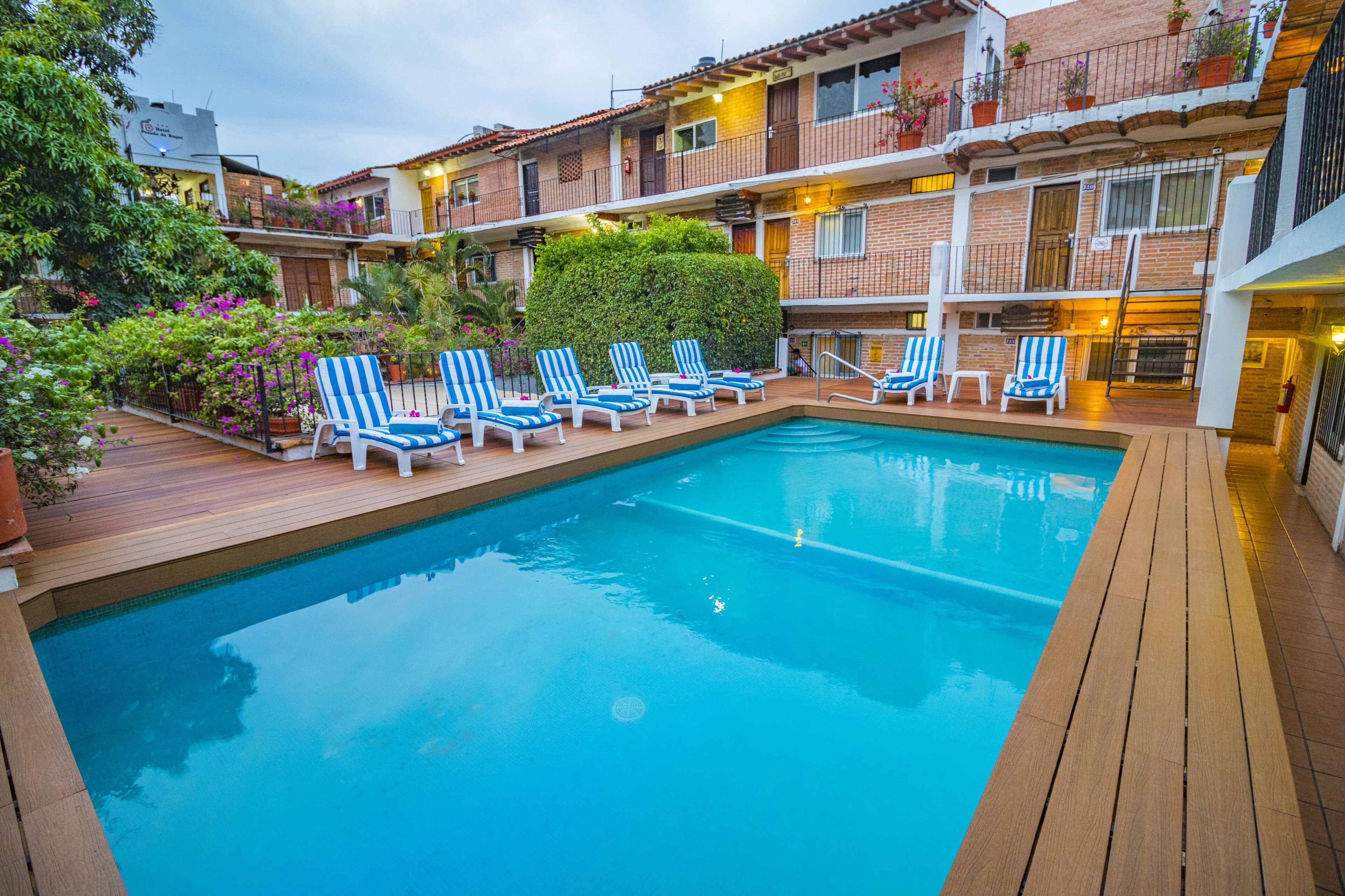 A swimming pool with lounge chairs, surrounded by a wooden deck and a brick building with balconies.