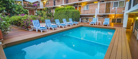 Une piscine entourée de chaises longues, d’une terrasse en bois et d’un bâtiment en briques doté de balcons.