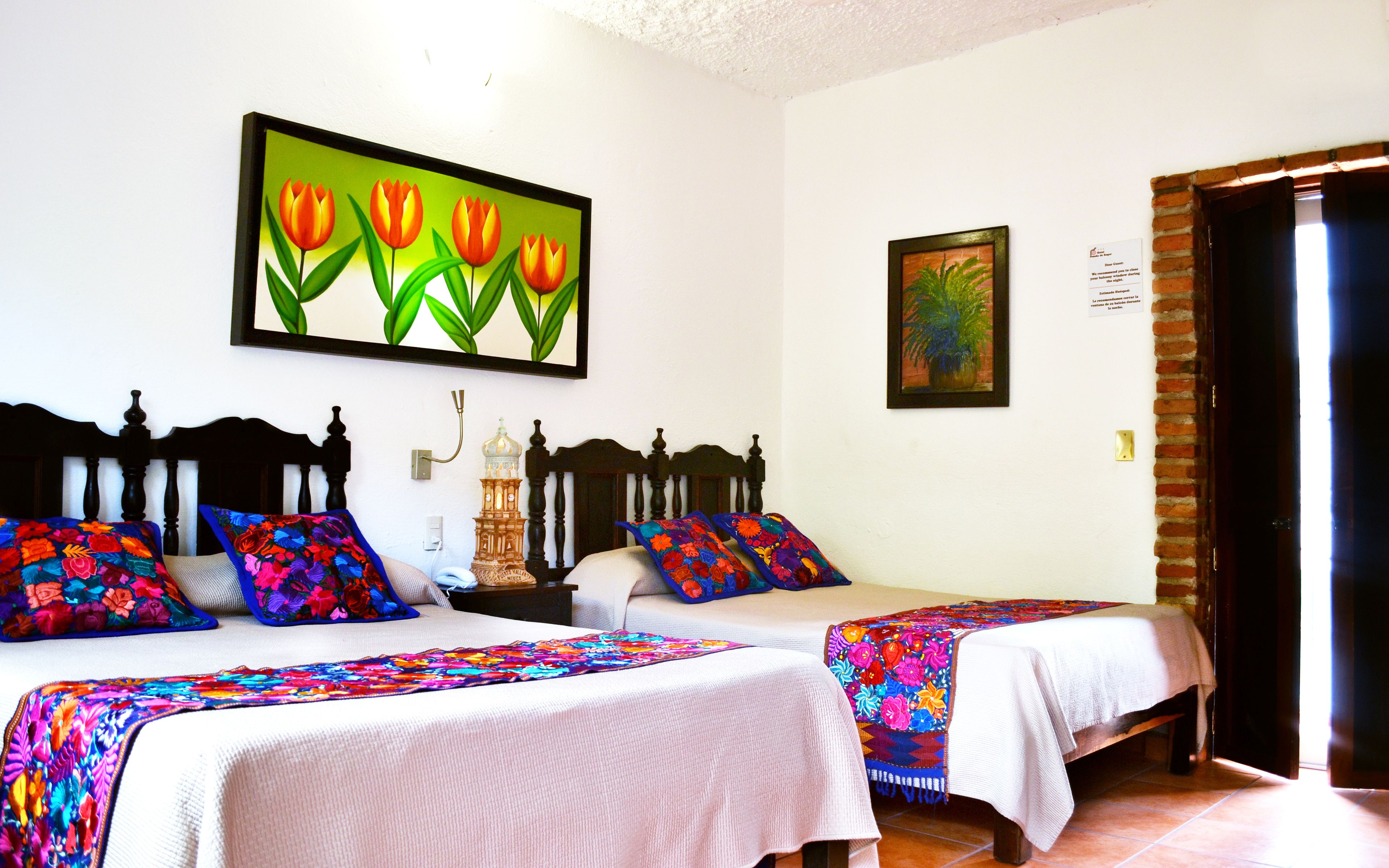 A room with two beds, each with a colorful quilt, a wall-mounted painting of flowers, and a small table with a lamp.