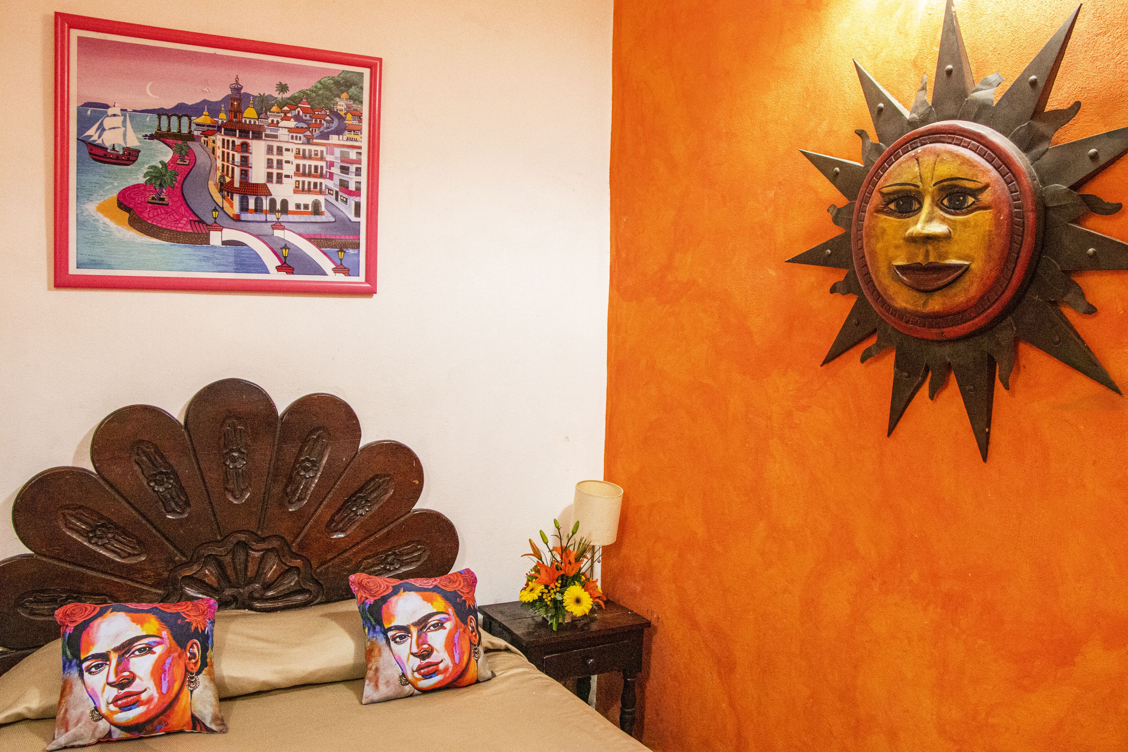 A bed with a headboard, a framed painting, a decorative sun, and a small table with a lamp and flowers.