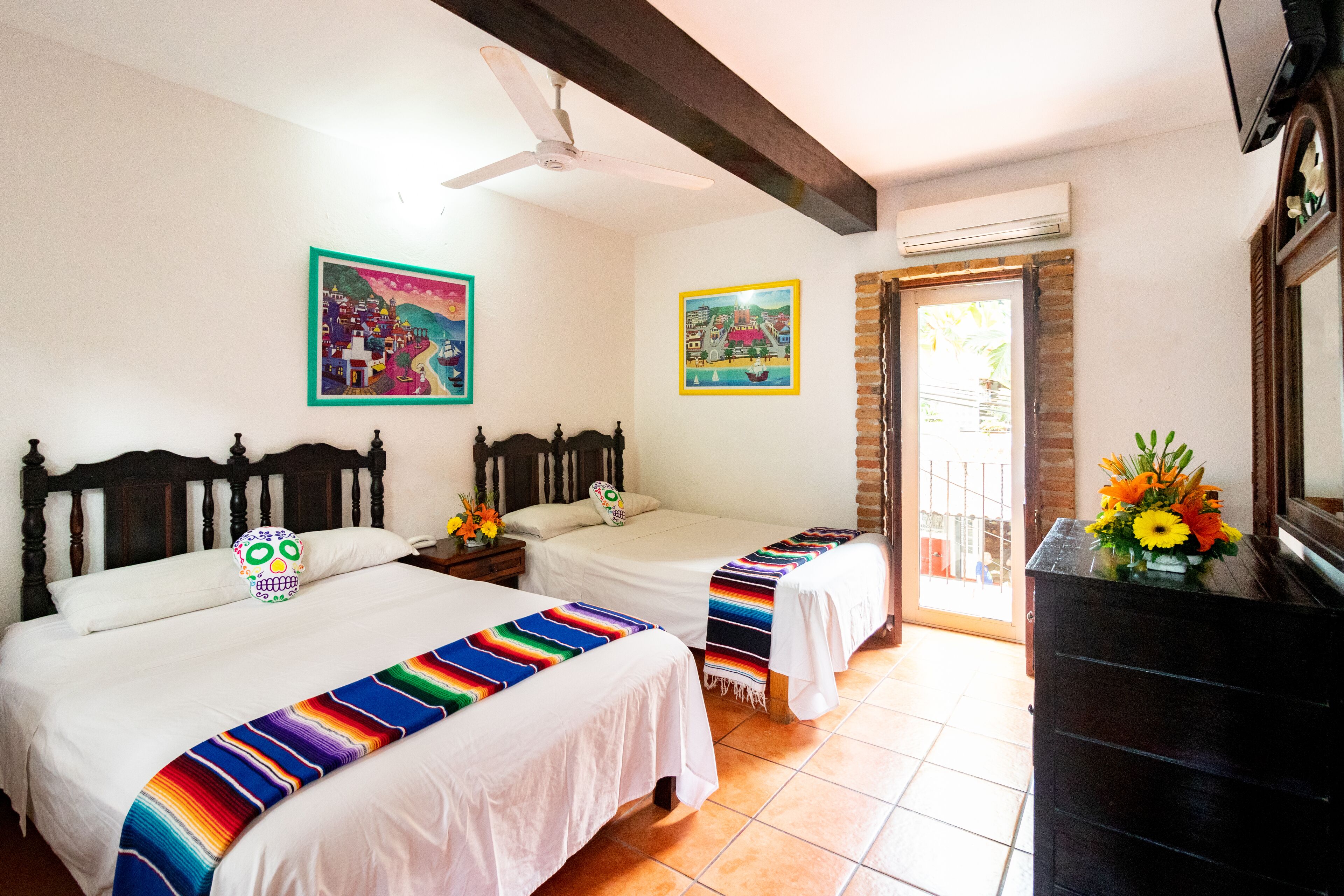 A hotel room with two beds, a ceiling fan, a TV, and artwork on the walls.
