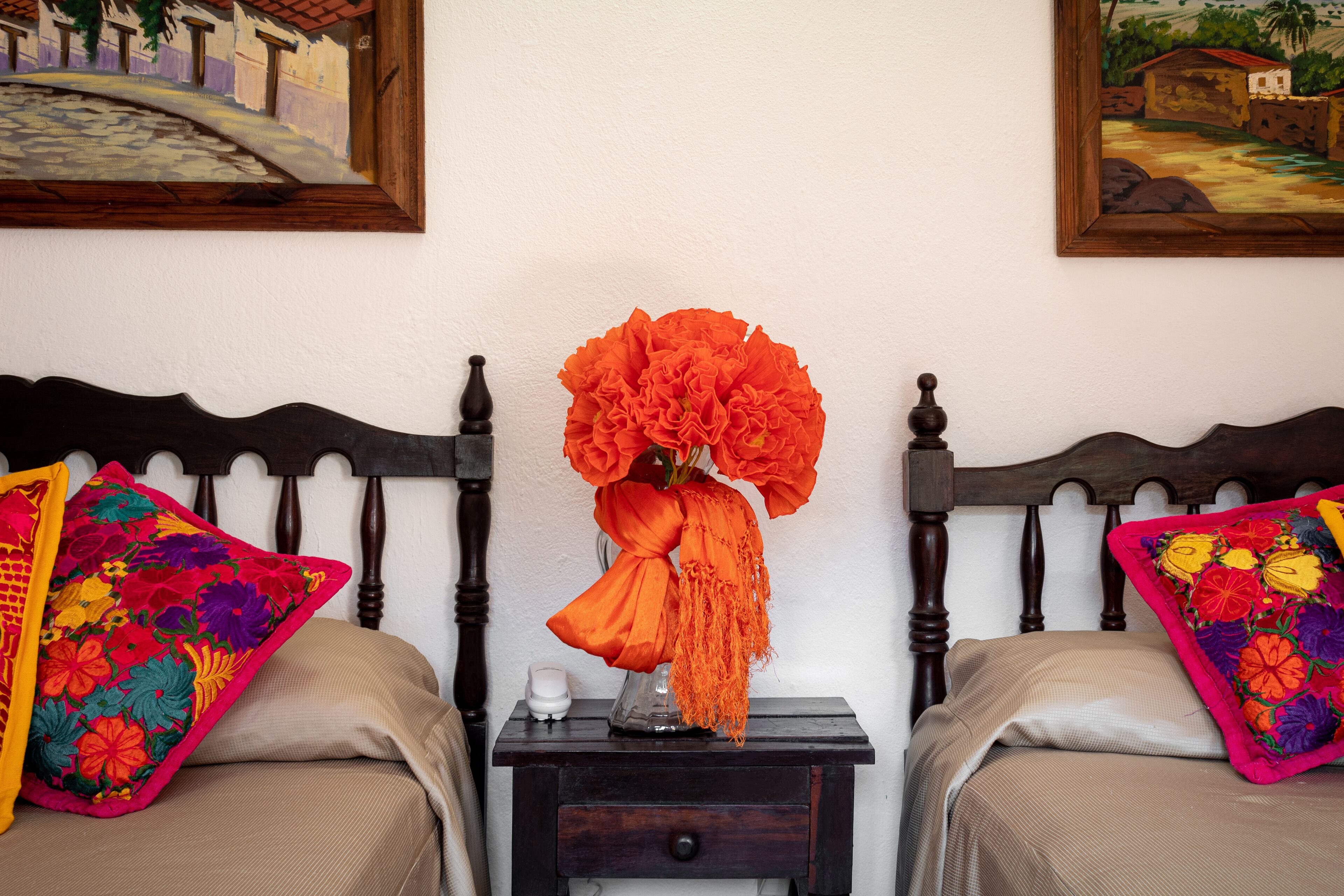 Una cama con cabecera de madera, una mesita de noche con un florero de flores naranjas y almohadones bordados de colores.