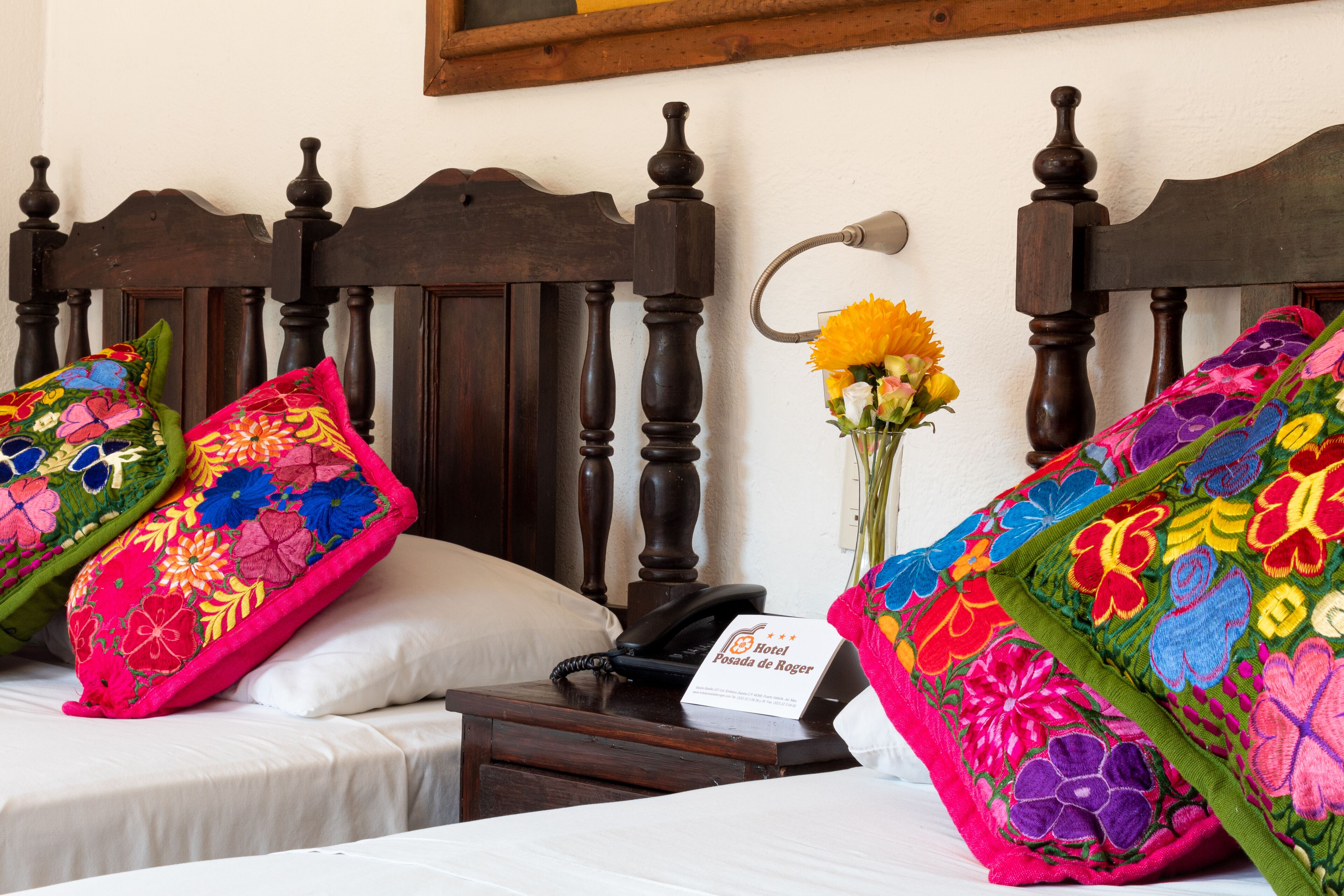 A hotel room with two beds, each with colorful embroidered pillows, a bedside table with a phone and a sign, and a vase with flowers.