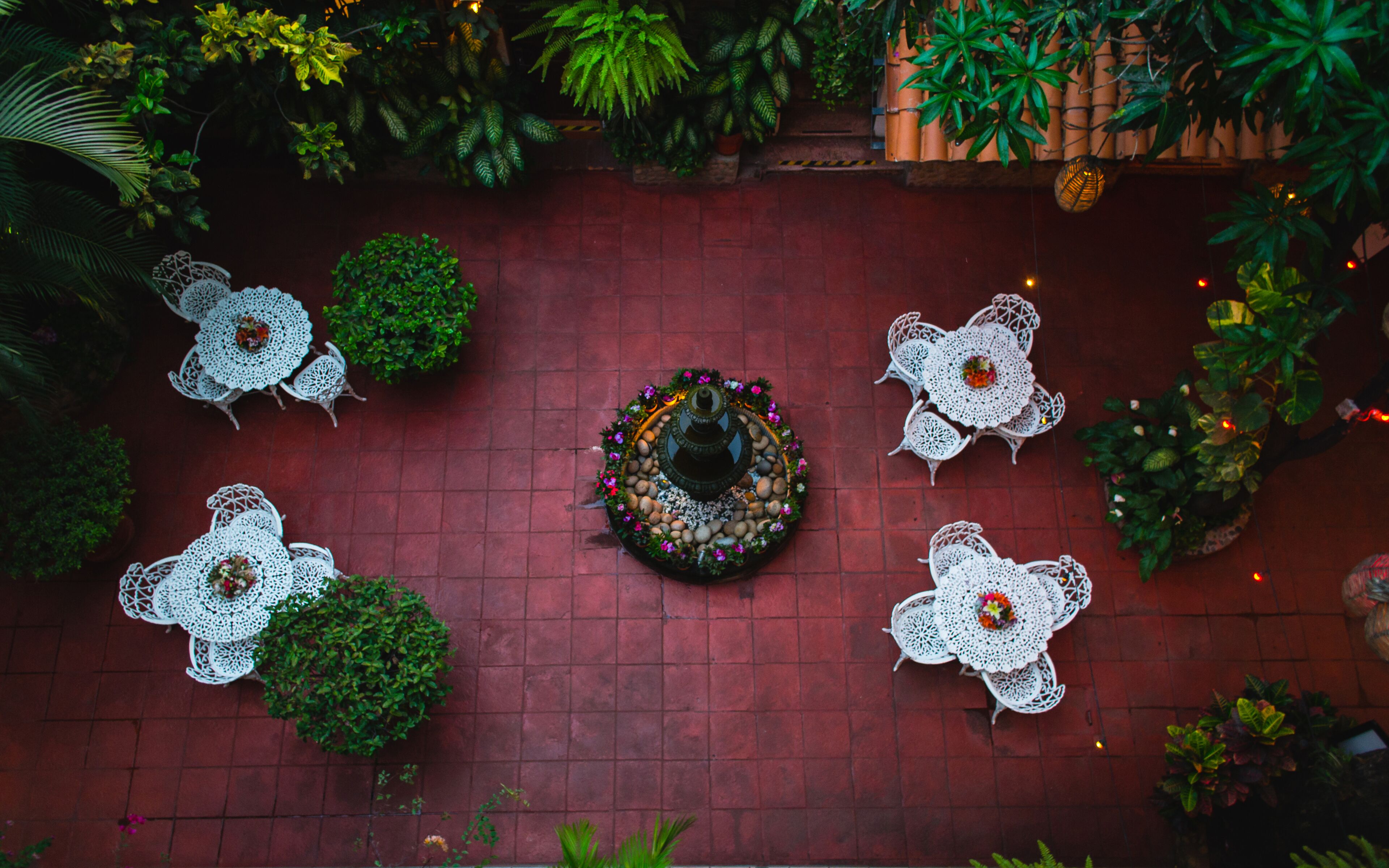 A top-down view of a courtyard with a central water feature surrounded by white wrought iron tables and chairs, and various potted plants and greenery around the perimeter.