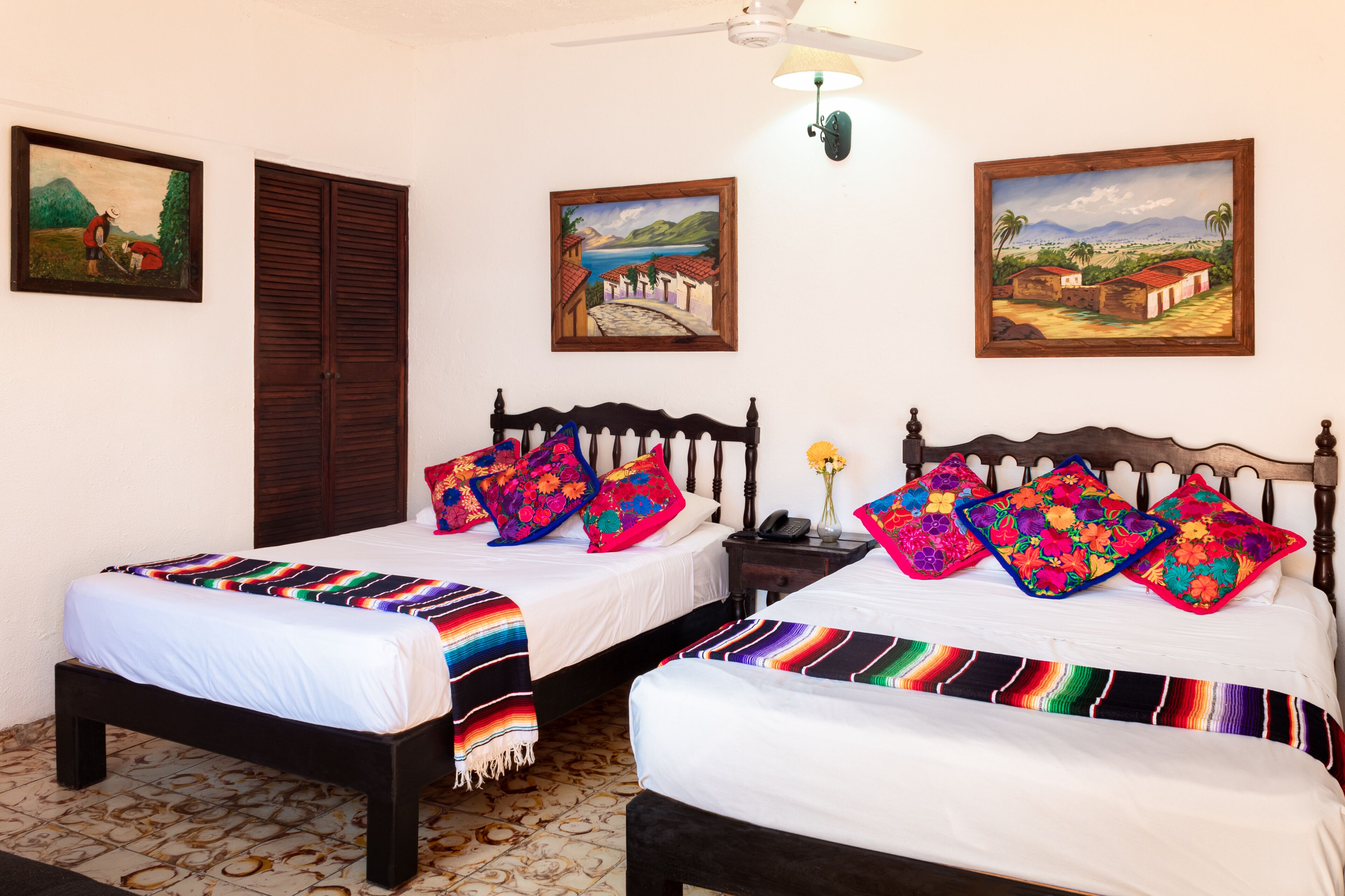 A room with two beds, each with colorful pillows and a striped blanket. There are paintings on the walls and a ceiling fan.