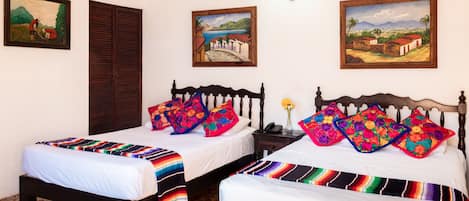 A room with two beds, each with colorful pillows and a striped blanket. There are paintings on the walls and a ceiling fan.